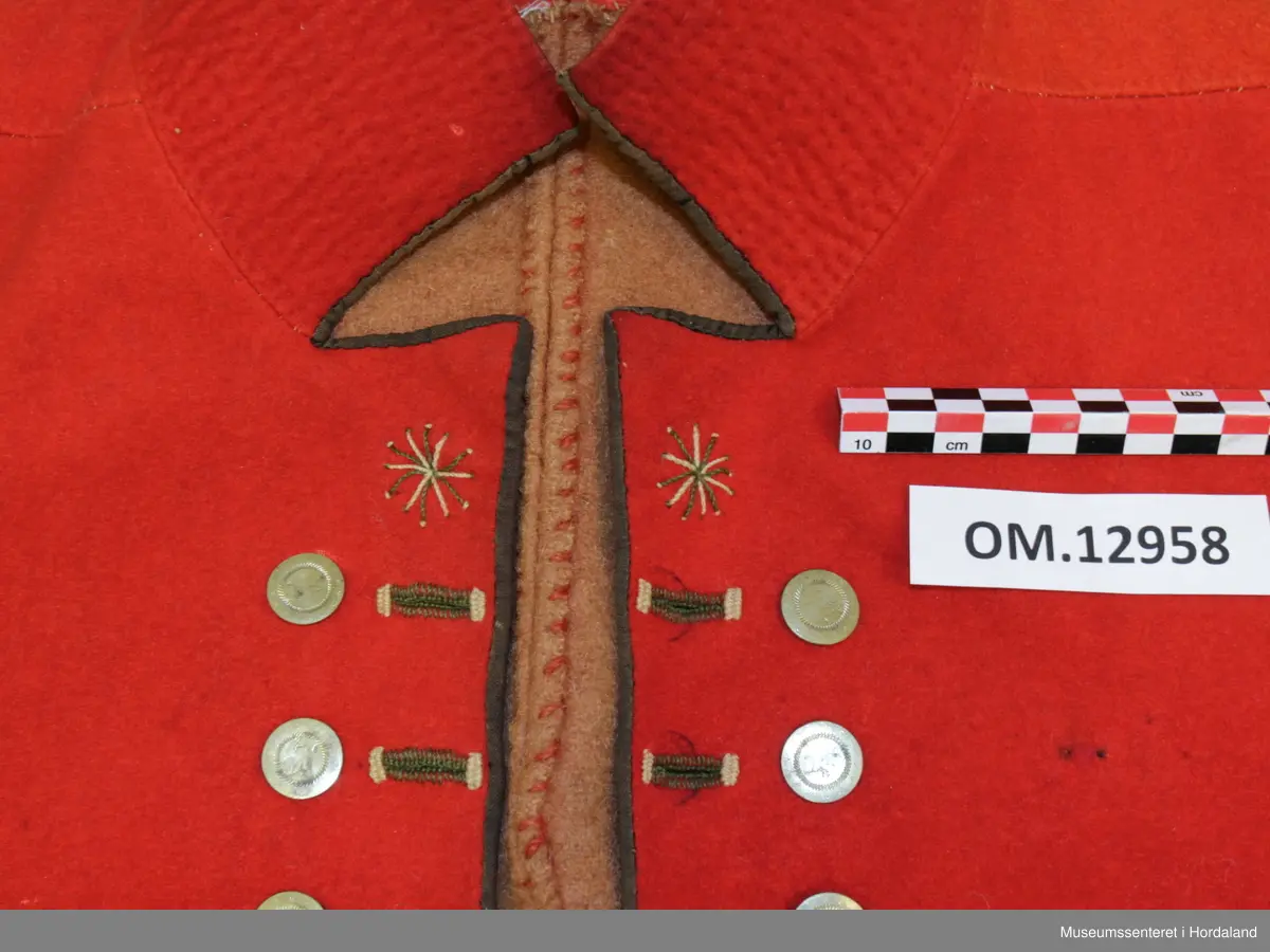 Dobbelknappa, raud mannsvest i ull med høg, raud krage. To lommer. Ei innfelt, raud stoffstripe oppå kvar skulder. Vesten er kanta med brunt silkeband framme i opninga og på lommene. Ti, tynne gullfarga knappar med knapphol på kvar side av opninga. Knapphola er sydde med ein brungrønfarga tråd, og med ein gulfarga- i endane. Lite, brodert stjerne-/blomstermotiv på kvar side oppe i spissane ved halsen, med dei same to trådfargane. Den høgreiste kragen er dobbel og er stiva av med tette horisontale saumar frå baksida. Tjukt, brunt vadmålsstoff i rygg. Framstykka er fora med bomullstoff, men det er linstoff i lommene. I spissane ved halsen er det på baksida sydd inn ein liten bit av eit mønstra bomullstoff i raudt, blått og ubleika farge. Heile vesten er handsauma.