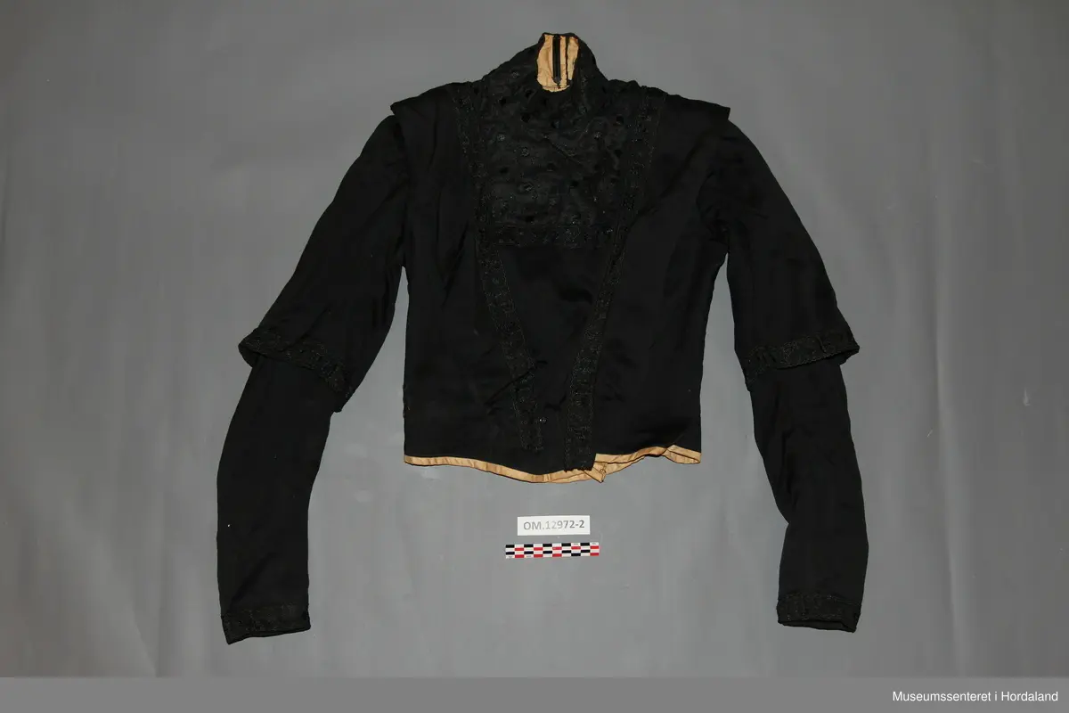 Svart brurekjole i to deler i ullsateng. Sidt skjørt og langarma bluse. Begge delane er fora med bomullsstoff, og er pynta med eit svart band. Blusen har i tillegg eit blondestoff i bringa, på kragen, og øvst i nakken. Kragen er høg og går heilt rundt. Han har støttespiler, og patentar til attlat. Blusen har først eit attlat med hemper på forstoffet i venstre framstykke som skal festast under høgre framstykke. Høgre framstykke med frontstykke skal så festast utanpå det venstre framstykket med hekter. Til slutt festast ein tamp eller sele over opninga med patentar. Denne går over skuldra frå nedst på ryggen til nedst på framstykket. På høgre framstykke er denne sydd fast. Blusen er kort, men er litt sidare midt framme og midt bak. Armane er rynka oppe ved skuldra og er korte, men med lang arm inni. Der dei lange armane er dekte av kortarmane, er det berre forstoff.