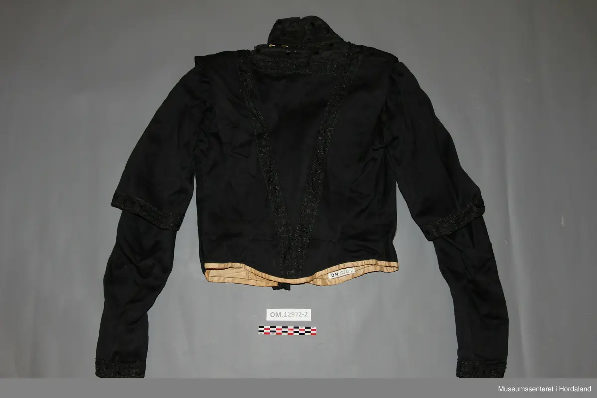 Svart brurekjole i to deler i ullsateng. Sidt skjørt og langarma bluse. Begge delane er fora med bomullsstoff, og er pynta med eit svart band. Blusen har i tillegg eit blondestoff i bringa, på kragen, og øvst i nakken. Kragen er høg og går heilt rundt. Han har støttespiler, og patentar til attlat. Blusen har først eit attlat med hemper på forstoffet i venstre framstykke som skal festast under høgre framstykke. Høgre framstykke med frontstykke skal så festast utanpå det venstre framstykket med hekter. Til slutt festast ein tamp eller sele over opninga med patentar. Denne går over skuldra frå nedst på ryggen til nedst på framstykket. På høgre framstykke er denne sydd fast. Blusen er kort, men er litt sidare midt framme og midt bak. Armane er rynka oppe ved skuldra og er korte, men med lang arm inni. Der dei lange armane er dekte av kortarmane, er det berre forstoff.