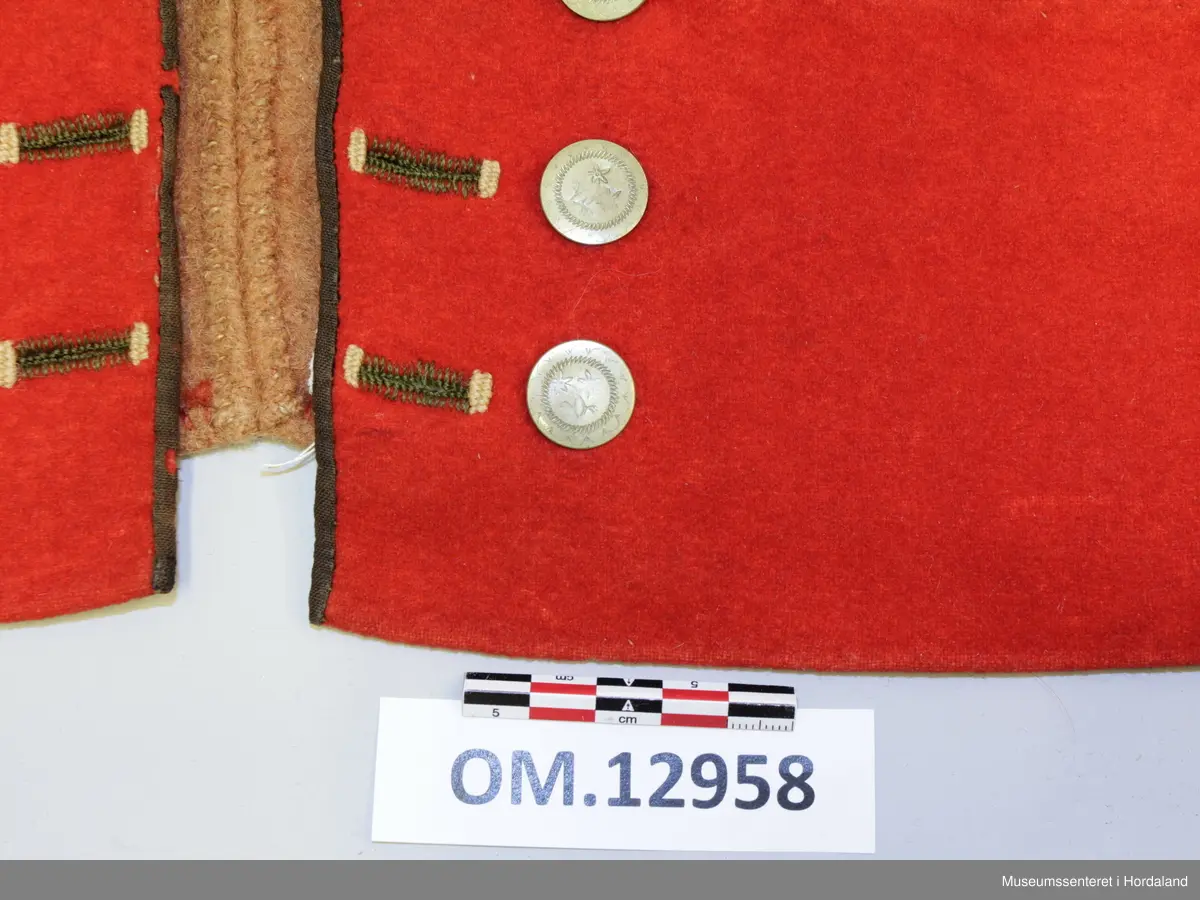 Dobbelknappa, raud mannsvest i ull med høg, raud krage. To lommer. Ei innfelt, raud stoffstripe oppå kvar skulder. Vesten er kanta med brunt silkeband framme i opninga og på lommene. Ti, tynne gullfarga knappar med knapphol på kvar side av opninga. Knapphola er sydde med ein brungrønfarga tråd, og med ein gulfarga- i endane. Lite, brodert stjerne-/blomstermotiv på kvar side oppe i spissane ved halsen, med dei same to trådfargane. Den høgreiste kragen er dobbel og er stiva av med tette horisontale saumar frå baksida. Tjukt, brunt vadmålsstoff i rygg. Framstykka er fora med bomullstoff, men det er linstoff i lommene. I spissane ved halsen er det på baksida sydd inn ein liten bit av eit mønstra bomullstoff i raudt, blått og ubleika farge. Heile vesten er handsauma.