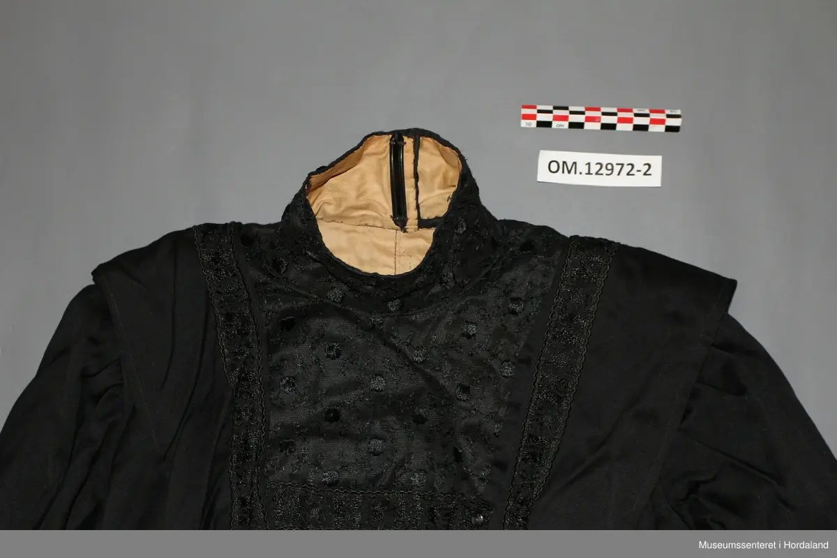 Svart brurekjole i to deler i ullsateng. Sidt skjørt og langarma bluse. Begge delane er fora med bomullsstoff, og er pynta med eit svart band. Blusen har i tillegg eit blondestoff i bringa, på kragen, og øvst i nakken. Kragen er høg og går heilt rundt. Han har støttespiler, og patentar til attlat. Blusen har først eit attlat med hemper på forstoffet i venstre framstykke som skal festast under høgre framstykke. Høgre framstykke med frontstykke skal så festast utanpå det venstre framstykket med hekter. Til slutt festast ein tamp eller sele over opninga med patentar. Denne går over skuldra frå nedst på ryggen til nedst på framstykket. På høgre framstykke er denne sydd fast. Blusen er kort, men er litt sidare midt framme og midt bak. Armane er rynka oppe ved skuldra og er korte, men med lang arm inni. Der dei lange armane er dekte av kortarmane, er det berre forstoff.