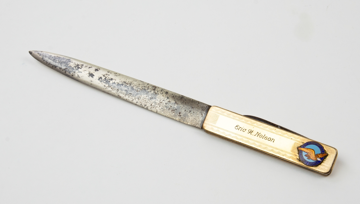 Papperskniv med pennkniv i skaftet. På skaftet inskriptionen Eric H Nelson samt ett emblem med texten "pratt and whitney U.S.A Dependable Engines" på mörkblå botten runt en ljusblå rundel med en flygande fågel i guldinläggning.  Tillverkare Remington U M G USA.