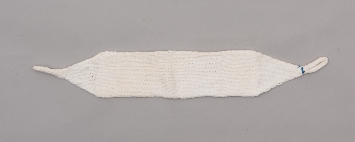 Håndstrikket hvitt bind uten åpning, med stropper i begge ender til festing. Merket med litt blått garn ved ene stropp, for å skille det fra andre bind.