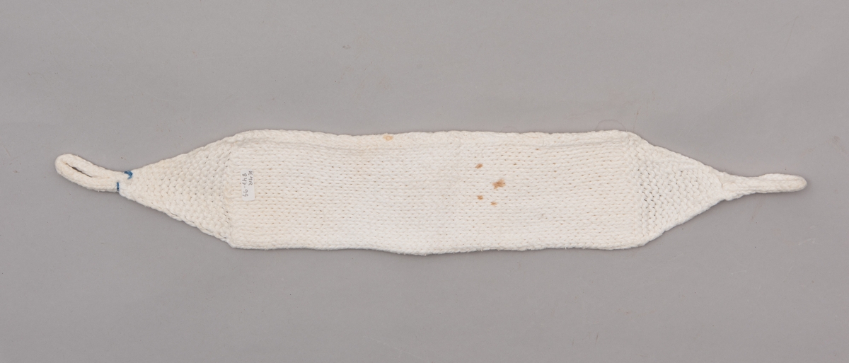 Håndstrikket hvitt bind uten åpning, med stropper i begge ender til festing. Merket med litt blått garn ved ene stropp, for å skille det fra andre bind.