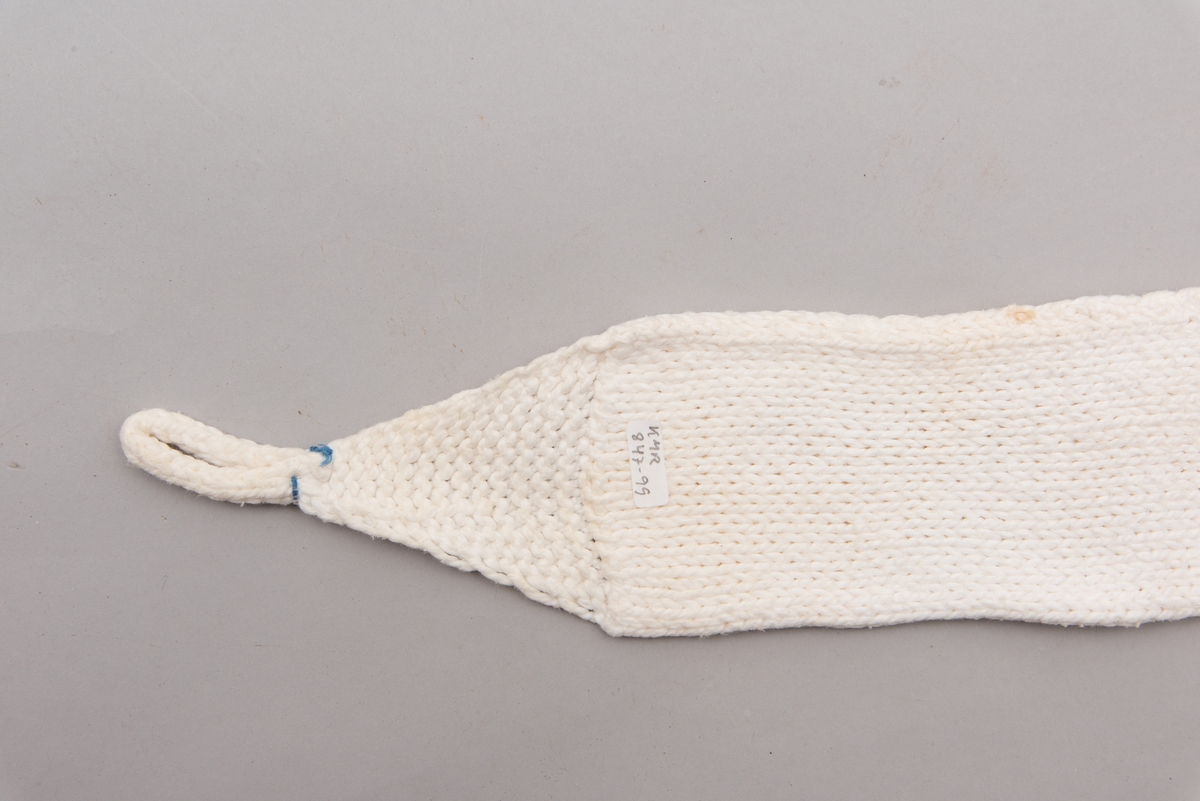 Håndstrikket hvitt bind uten åpning, med stropper i begge ender til festing. Merket med litt blått garn ved ene stropp, for å skille det fra andre bind.