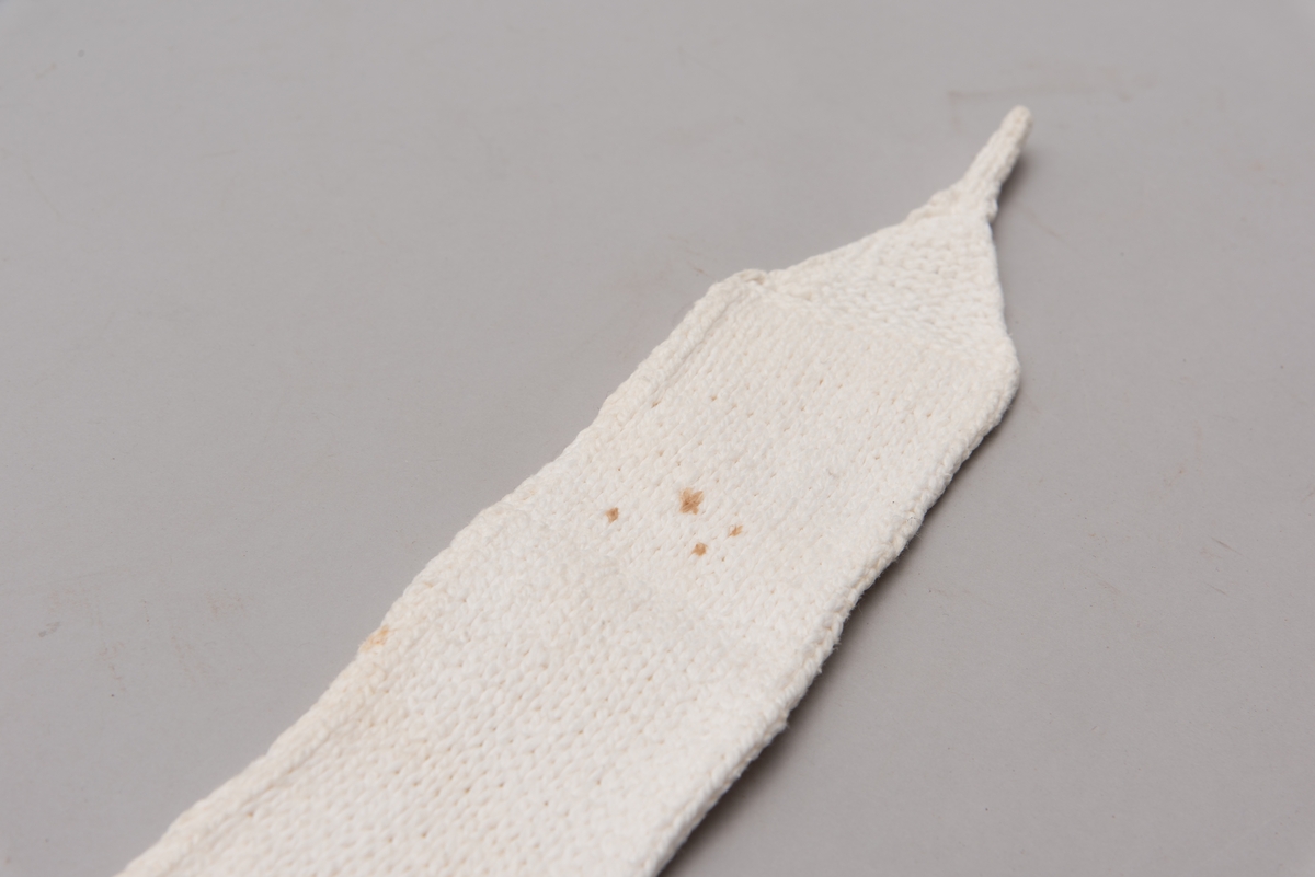 Håndstrikket hvitt bind uten åpning, med stropper i begge ender til festing. Merket med litt blått garn ved ene stropp, for å skille det fra andre bind.