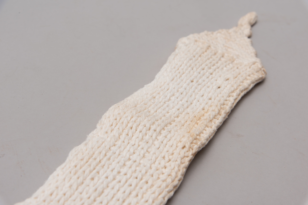 Håndstrikket hvitt bind med åpning, med stropper i begge ender til festing. Strikket i ribbestrikk men en rett og en vrang. Endekanter og stropper er heklet.