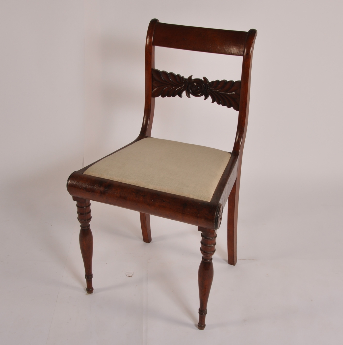 Stol i mahogny. Biedermeier. Lett konkav rygg, rett toppstykke, horisontaltstillt ryggbrett med sirkel og stiliserte blader. Sargen avsluttet halvsirkelformet foran med to dreide klosser. Sabelformede bakben i forlengelse av ryggen, dreide forben. Løst sete med salgjord, mangler trekk og har hvitt tynt bomullstrekk.