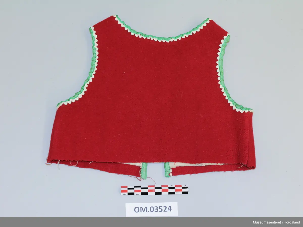 Raudt barneliv med lysegrøne silkeband til kantband og kvite silkekrokalisser i bølgjeform innom. Rund halsringning som fylgjer kragebeinet. Opning framme, 2 hekter nede. Fora.