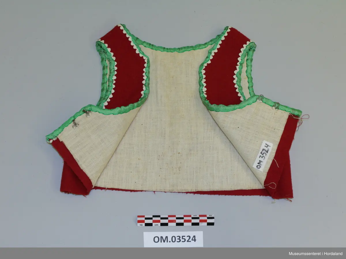 Raudt barneliv med lysegrøne silkeband til kantband og kvite silkekrokalisser i bølgjeform innom. Rund halsringning som fylgjer kragebeinet. Opning framme, 2 hekter nede. Fora.