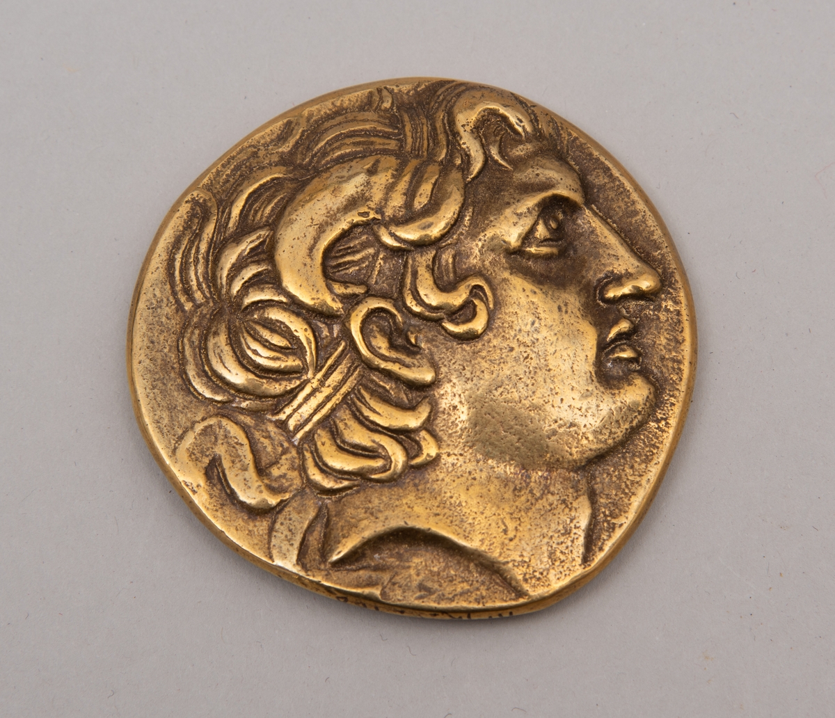 Brevpresse i bronse, med eske og informasjonsfolder. Motiv (baksiden) av gudinnen Athene på tronen som holder Nike. På forsiden portrett av Aleksander den store. Brevpressen er fra museumsbutikken til Benaki Museum i Athen. På informasjonstekst i esken står: Bronze replica. Gold stater of Lysimachous of Thrace (306-281 BC).