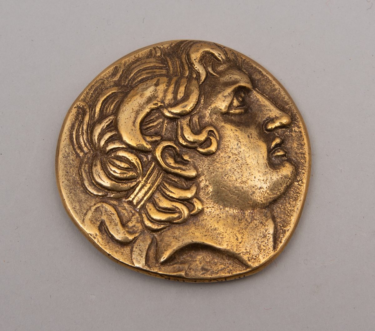 Brevpresse i bronse, med eske og informasjonsfolder. Motiv (baksiden) av gudinnen Athene på tronen som holder Nike. På forsiden portrett av Aleksander den store. Brevpressen er fra museumsbutikken til Benaki Museum i Athen. På informasjonstekst i esken står: Bronze replica. Gold stater of Lysimachous of Thrace (306-281 BC).