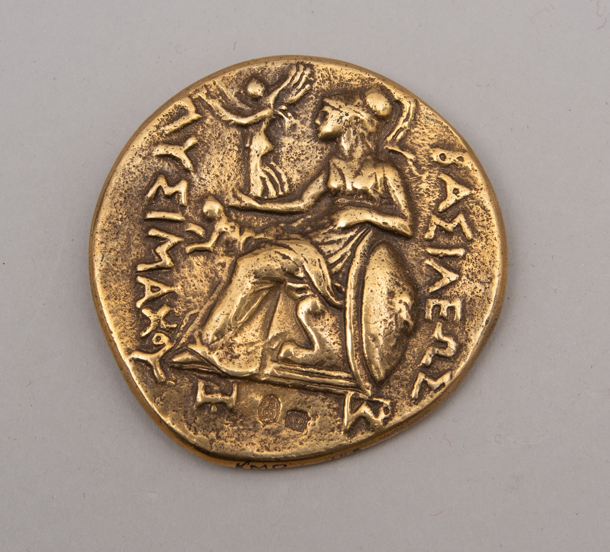 Brevpresse i bronse, med eske og informasjonsfolder. Motiv (baksiden) av gudinnen Athene på tronen som holder Nike. På forsiden portrett av Aleksander den store. Brevpressen er fra museumsbutikken til Benaki Museum i Athen. På informasjonstekst i esken står: Bronze replica. Gold stater of Lysimachous of Thrace (306-281 BC).