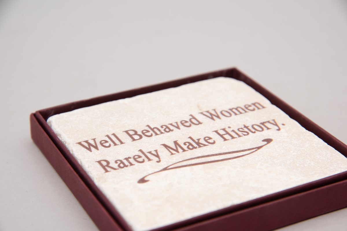 Marmorflis og souvenir fra Newsport Mansions med påført slagord "Well Behaved Women Rarely Make History". Tilhørende eske og informasjonsfolder. Mottoet påført marmorflisa er i ettertida blitt brukt til å beskrive Alva Vanderbilt Belmont (1853-1933) og andre kvinner som kjempet for kvinners stemmerett på begynnelsen av 1900-tallet i USA under slagordet "Votes for Women". Selve "Votes for Women"-mottoet ble brukt i 1909 på et berømt "open house" stemmerettsmøte holdt på Marble House, Newport, Rhode Island".