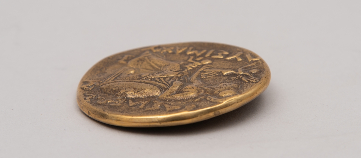 Brevpresse i bronse, med eske og informasjonsfolder. Motiv (baksiden) av gudinnen Athene på tronen som holder Nike. På forsiden portrett av Aleksander den store. Brevpressen er fra museumsbutikken til Benaki Museum i Athen. På informasjonstekst i esken står: Bronze replica. Gold stater of Lysimachous of Thrace (306-281 BC).