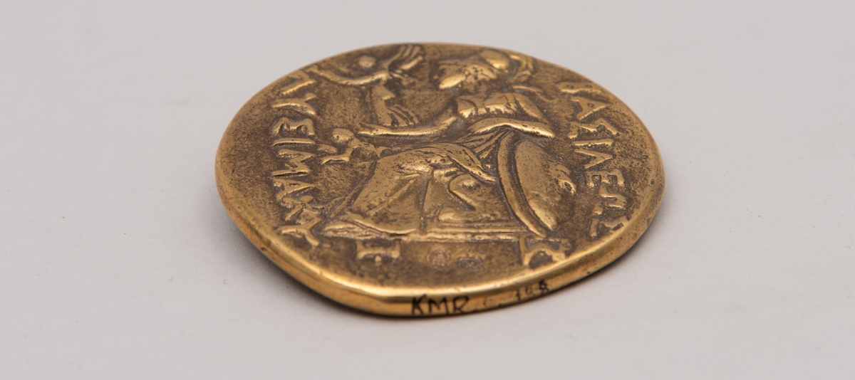 Brevpresse i bronse, med eske og informasjonsfolder. Motiv (baksiden) av gudinnen Athene på tronen som holder Nike. På forsiden portrett av Aleksander den store. Brevpressen er fra museumsbutikken til Benaki Museum i Athen. På informasjonstekst i esken står: Bronze replica. Gold stater of Lysimachous of Thrace (306-281 BC).
