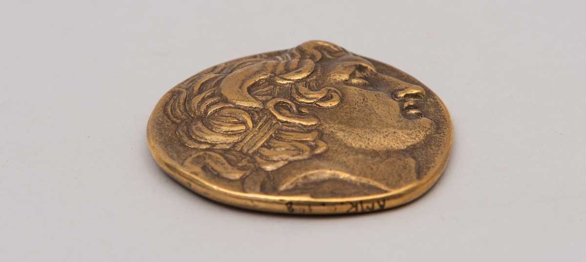 Brevpresse i bronse, med eske og informasjonsfolder. Motiv (baksiden) av gudinnen Athene på tronen som holder Nike. På forsiden portrett av Aleksander den store. Brevpressen er fra museumsbutikken til Benaki Museum i Athen. På informasjonstekst i esken står: Bronze replica. Gold stater of Lysimachous of Thrace (306-281 BC).