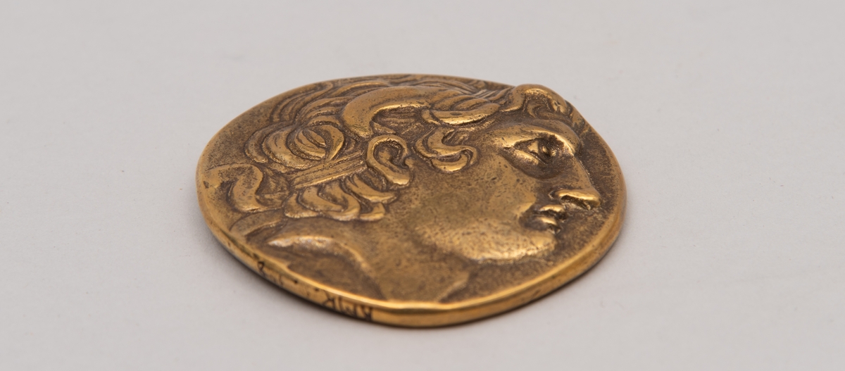 Brevpresse i bronse, med eske og informasjonsfolder. Motiv (baksiden) av gudinnen Athene på tronen som holder Nike. På forsiden portrett av Aleksander den store. Brevpressen er fra museumsbutikken til Benaki Museum i Athen. På informasjonstekst i esken står: Bronze replica. Gold stater of Lysimachous of Thrace (306-281 BC).
