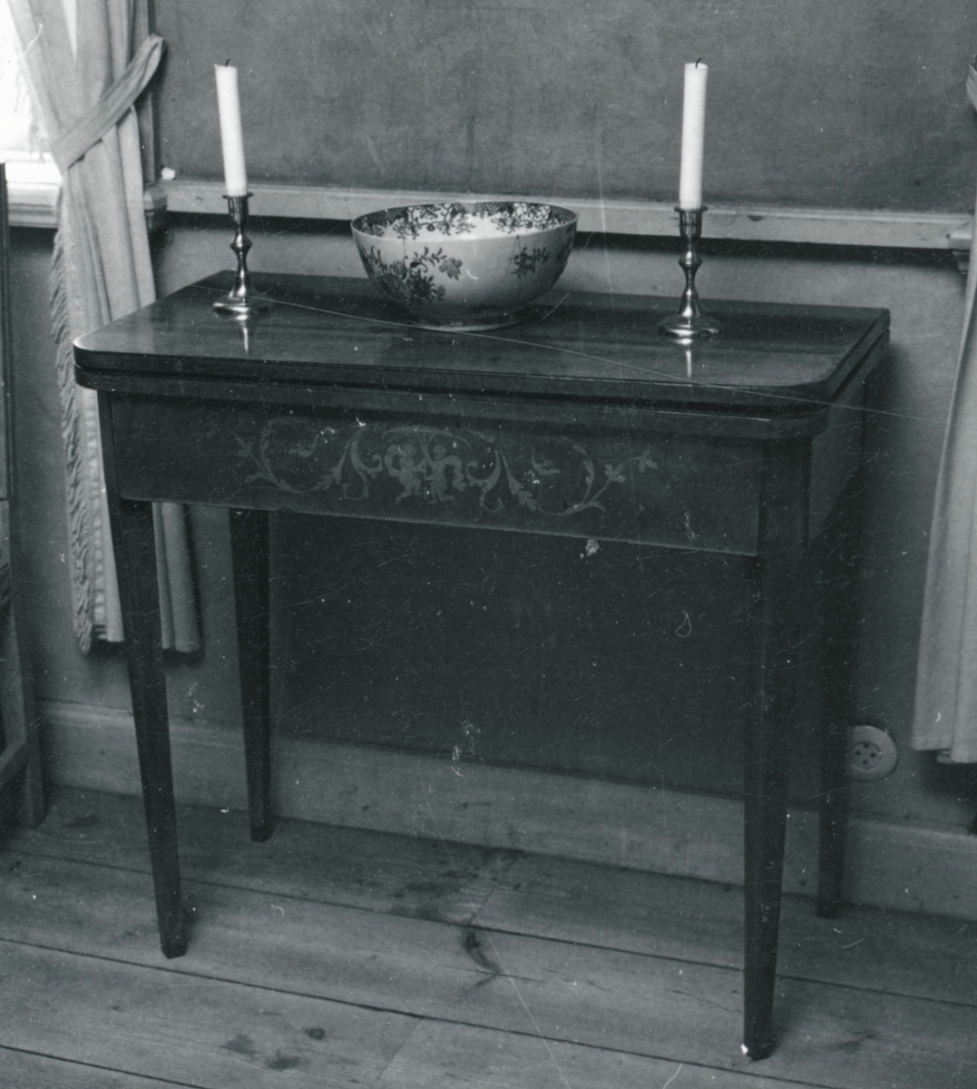 Spelbord av mahogny. På framsidan av sargen dekorationer i intarsia. Skivan, nedfällbar, är liksidig med avrundade hörn. Raka, nedåt avsmalnande ben. Se 1M16-86191.