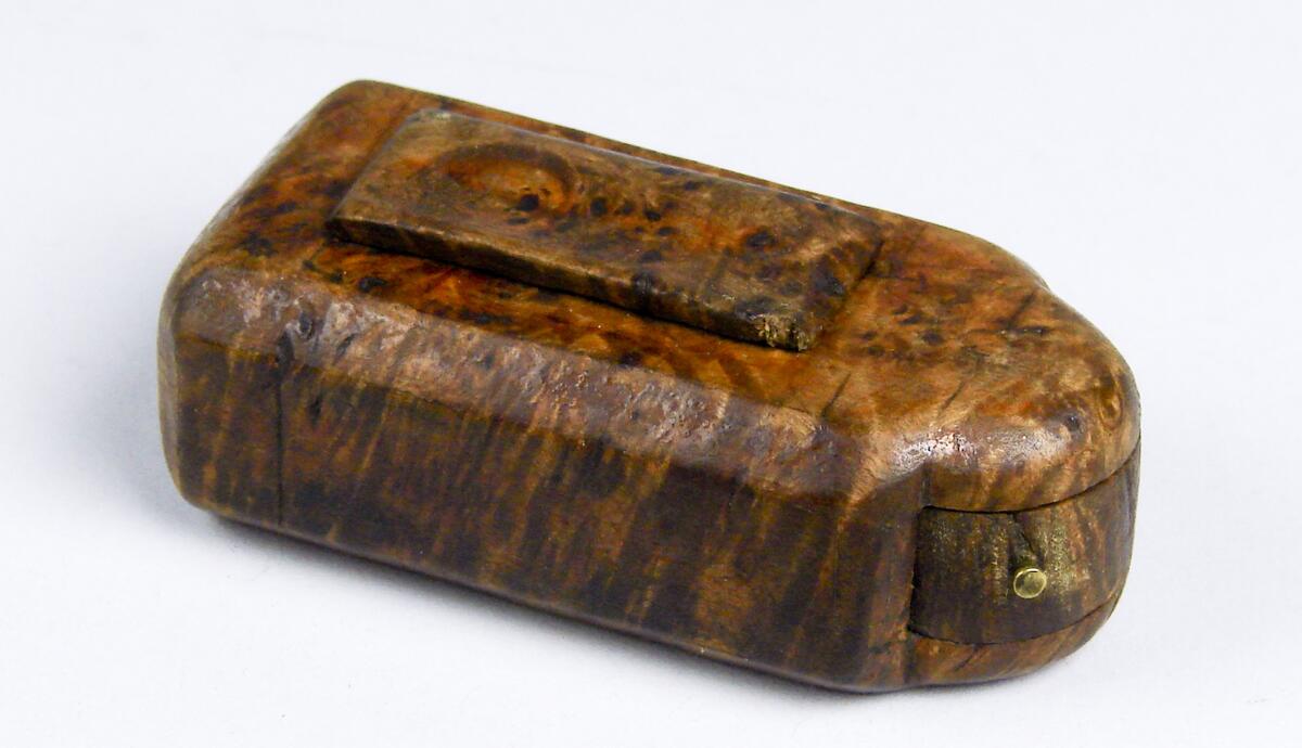 Snusdosa, av masur. Rektangulär med ena kortsidan kraftigt utsvängd. Denna sida med vridbart hjul. Litet lock, märkt 1802. Ramsele socken, Ångermanland. (Ur lappkatalogen).