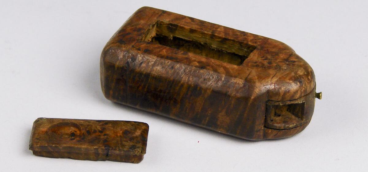 Snusdosa, av masur. Rektangulär med ena kortsidan kraftigt utsvängd. Denna sida med vridbart hjul. Litet lock, märkt 1802. Ramsele socken, Ångermanland. (Ur lappkatalogen).