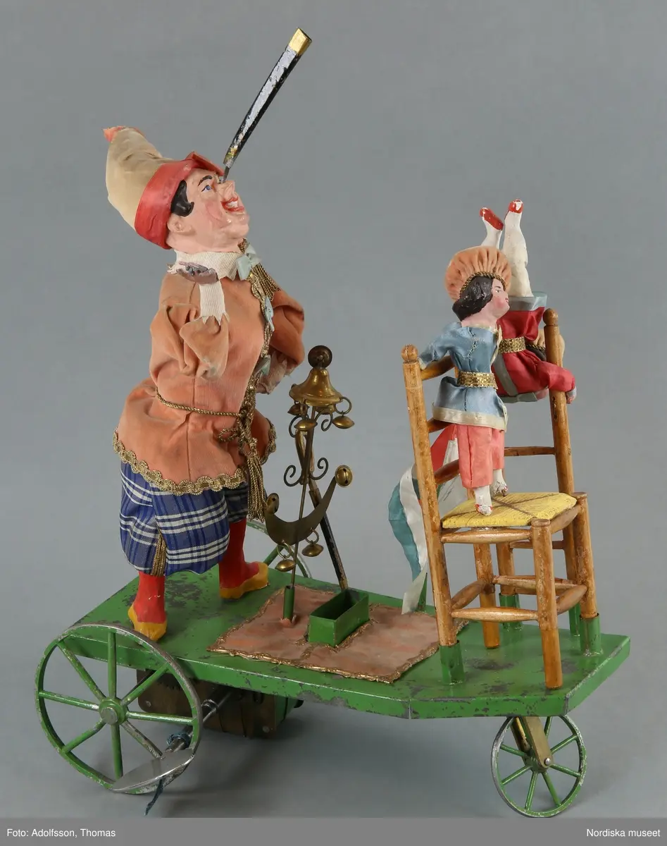 Inventering Sesam 1996-1999:
a+) Mekanisk leksak L 28 cm, B 12,5 cm, H) 45,5 cm
b) Nyckel L 10 cm
c) Originalförpackning L 37 cm, B 17 cm, H 35 cm
a+) Equilibriste ¿ Mecanique (mekanisk balanskonstnär) bestående av clown av massa (papier mach¿?), med rosa sammetsjacka och blårutiga byxor, stående på grönlackerad vagn av metall med tre hjul. På näsan har clownen fäste (spets) på vilken kan fästas. På vagnen olika fästen (bl a två svärdsskidor) för olika lösa attiraljer: Stege med docka i röd sidenjacka och beige huvudbonad, stol (delar saknas) av trä med gul sits och stående på sitsen liten docka med blå jacka, tvådelad flaggstång med grön/vit och röd/vit tvåuddiga flaggor, metallten med klocka och halvmåne i gulmetall med bjällor, Svärd av gulmetall (har ursprungligen trol. funnits två svärd)
Undertill på vagnen finns ett fjäderverk som driver vagnen frammåt och får fästet/spetsen på clownens näsa att snurra. Enligt äldre kat.kort driver fjäderverket även speldosa (?). På spetsen på clownens näsa kan de olika lösa attiraljerna fästas.
b) Nyckel, av vitmetall, ovalt huvud utan hål.
c+) Originalförpackning, enkel, av obehandlat trä, Stor lucka framtill, lock ovanpå. Enligt äldre kat.kort skall det ursprungligen ha suttit ett reklamkort på locket, vilket det nu bara finns reser kvar av.
Har tillhört brukspatron Edv. Otto Benedicks (1818-1872), Gysinge, Gästrikland. 
Leif Wallin feb 1998