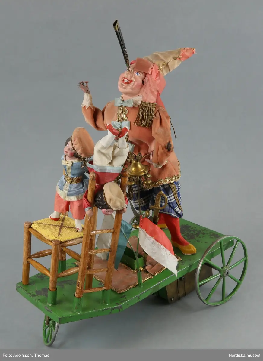 Inventering Sesam 1996-1999:
a+) Mekanisk leksak L 28 cm, B 12,5 cm, H) 45,5 cm
b) Nyckel L 10 cm
c) Originalförpackning L 37 cm, B 17 cm, H 35 cm
a+) Equilibriste ¿ Mecanique (mekanisk balanskonstnär) bestående av clown av massa (papier mach¿?), med rosa sammetsjacka och blårutiga byxor, stående på grönlackerad vagn av metall med tre hjul. På näsan har clownen fäste (spets) på vilken kan fästas. På vagnen olika fästen (bl a två svärdsskidor) för olika lösa attiraljer: Stege med docka i röd sidenjacka och beige huvudbonad, stol (delar saknas) av trä med gul sits och stående på sitsen liten docka med blå jacka, tvådelad flaggstång med grön/vit och röd/vit tvåuddiga flaggor, metallten med klocka och halvmåne i gulmetall med bjällor, Svärd av gulmetall (har ursprungligen trol. funnits två svärd)
Undertill på vagnen finns ett fjäderverk som driver vagnen frammåt och får fästet/spetsen på clownens näsa att snurra. Enligt äldre kat.kort driver fjäderverket även speldosa (?). På spetsen på clownens näsa kan de olika lösa attiraljerna fästas.
b) Nyckel, av vitmetall, ovalt huvud utan hål.
c+) Originalförpackning, enkel, av obehandlat trä, Stor lucka framtill, lock ovanpå. Enligt äldre kat.kort skall det ursprungligen ha suttit ett reklamkort på locket, vilket det nu bara finns reser kvar av.
Har tillhört brukspatron Edv. Otto Benedicks (1818-1872), Gysinge, Gästrikland. 
Leif Wallin feb 1998