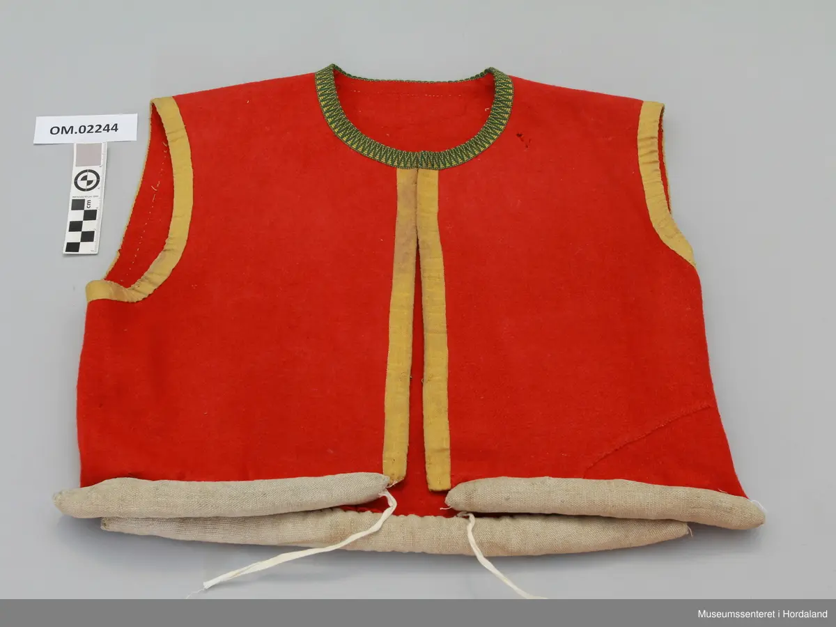 Raudt ull-liv utan fôr. Grøn og gul krokakvarde av silke langs halsopning. Gul silkekvarde langs frontopninga og rundt armhola. Ei hekte under bysten til attlat. Valk rundt kanten nede trekt med lin. I opninga heilt nede er det sydd på snorer av bomullsband (på valken). Livet er reparert nede på venstre framstykke med ein påsydd stoffbit. Handsauma.