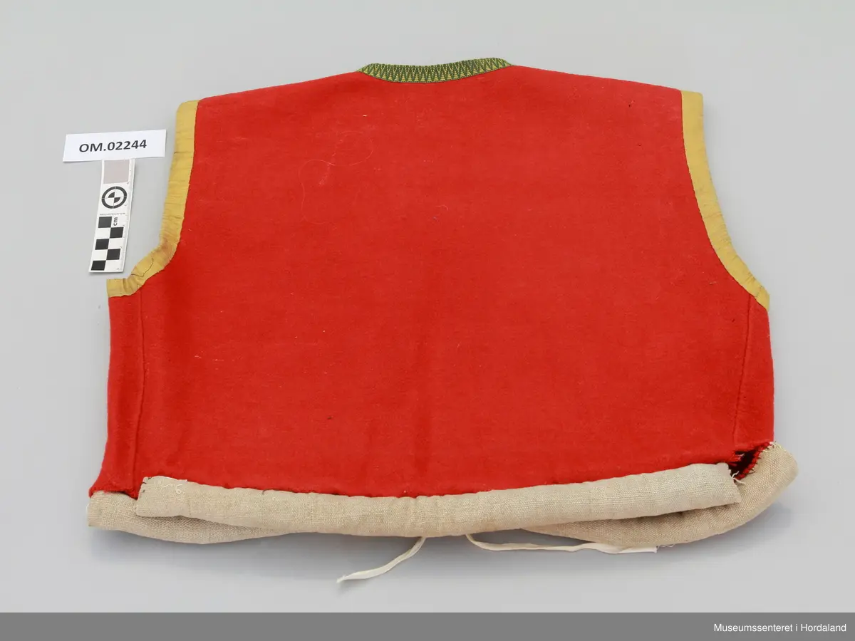 Raudt ull-liv utan fôr. Grøn og gul krokakvarde av silke langs halsopning. Gul silkekvarde langs frontopninga og rundt armhola. Ei hekte under bysten til attlat. Valk rundt kanten nede trekt med lin. I opninga heilt nede er det sydd på snorer av bomullsband (på valken). Livet er reparert nede på venstre framstykke med ein påsydd stoffbit. Handsauma.