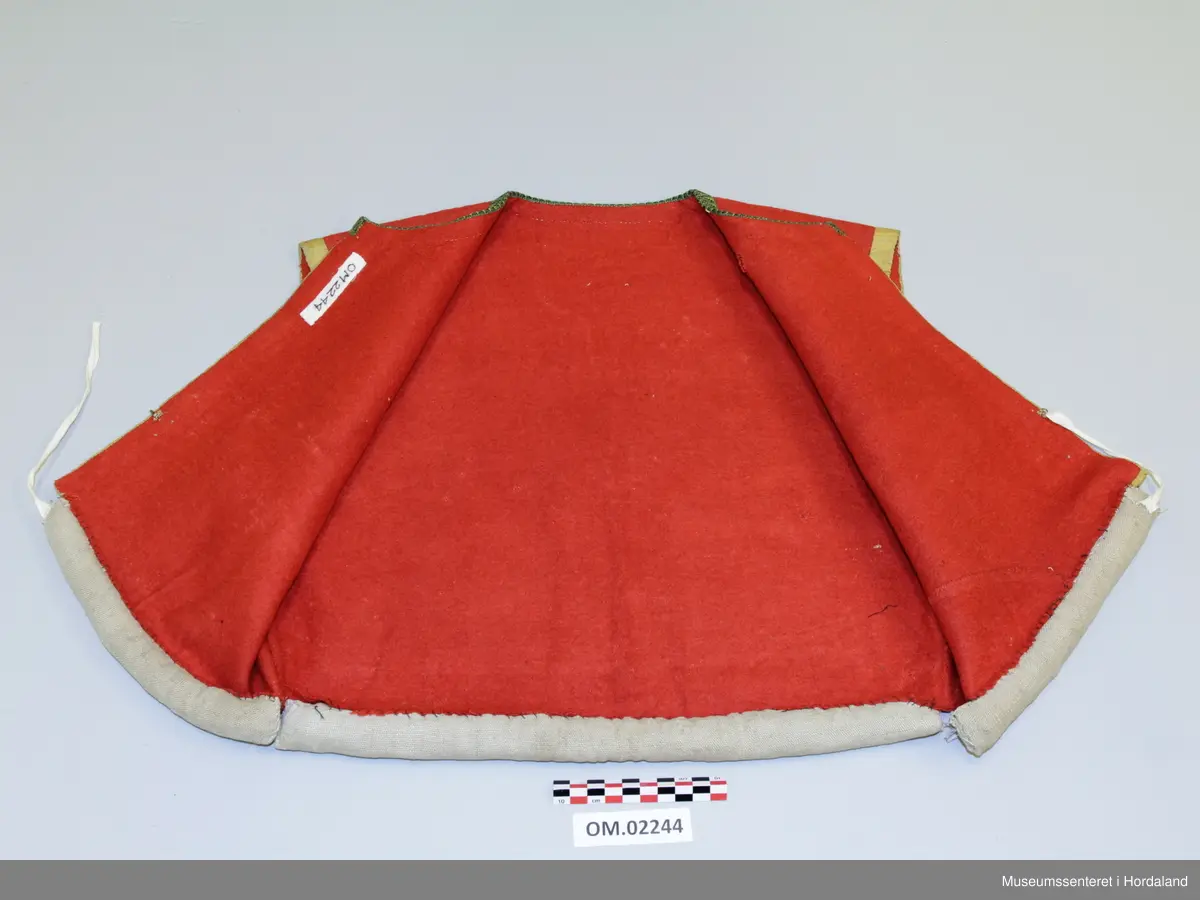 Raudt ull-liv utan fôr. Grøn og gul krokakvarde av silke langs halsopning. Gul silkekvarde langs frontopninga og rundt armhola. Ei hekte under bysten til attlat. Valk rundt kanten nede trekt med lin. I opninga heilt nede er det sydd på snorer av bomullsband (på valken). Livet er reparert nede på venstre framstykke med ein påsydd stoffbit. Handsauma.