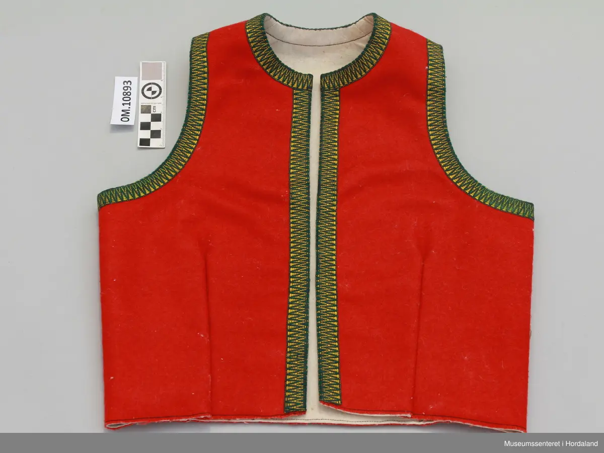 Raudt bunadsliv i ull, med grøn og gul krokakvarding i silke rundt armhola, hals og i front. Ikkje på skuldrar. Fora. Eitt innsnitt på kvart forstykke. Fire hekter til attlat framme.