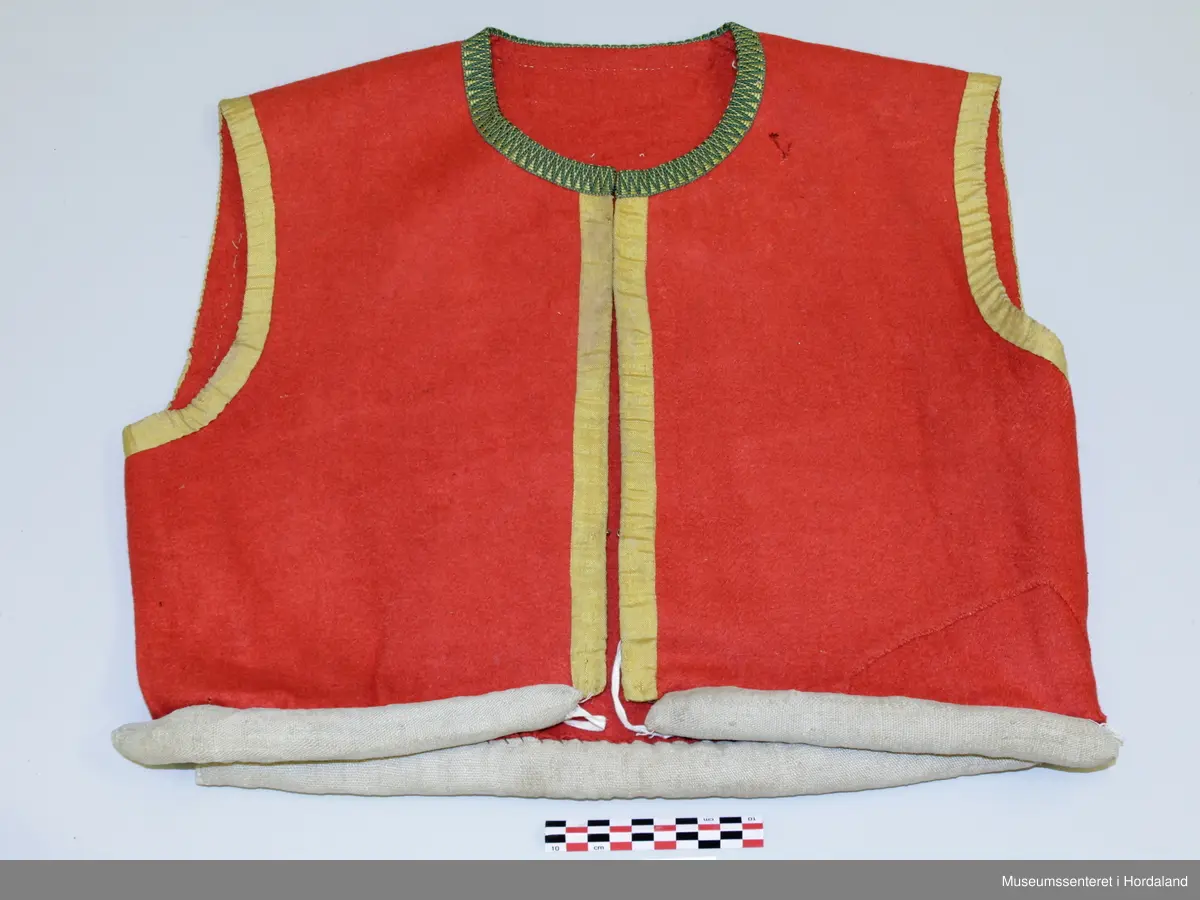 Raudt ull-liv utan fôr. Grøn og gul krokakvarde av silke langs halsopning. Gul silkekvarde langs frontopninga og rundt armhola. Ei hekte under bysten til attlat. Valk rundt kanten nede trekt med lin. I opninga heilt nede er det sydd på snorer av bomullsband (på valken). Livet er reparert nede på venstre framstykke med ein påsydd stoffbit. Handsauma.
