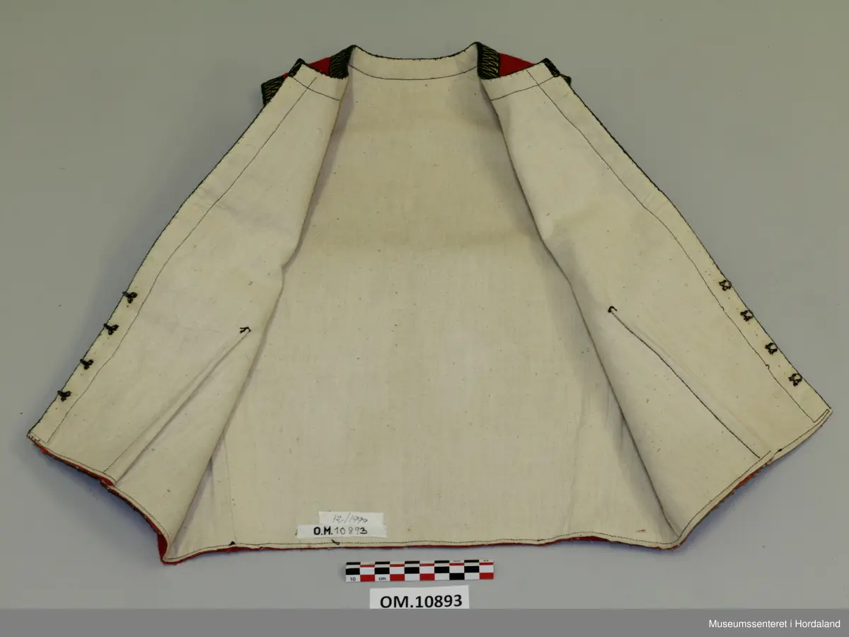 Raudt bunadsliv i ull, med grøn og gul krokakvarding i silke rundt armhola, hals og i front. Ikkje på skuldrar. Fora. Eitt innsnitt på kvart forstykke. Fire hekter til attlat framme.