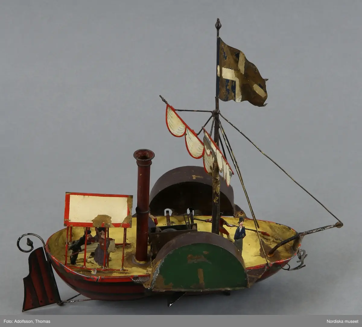 Katalogkort:
"Leksak, hjulångare med nyckel, mekanisk, bleckplåt, målad. 1800-talets mitt, Tyskland [med blyerts]. Spetsgattat fartyg, brunt skrov med svarta linjer; utanpåliggande roder, i fören ankare. gult däck; hög brun skorsten; mast med beslagna segel samt i toppen flagga. I aktern överbyggnad uppburen av tunnt rött galler. Vid friborden skovelhjul täckta av gröna och svarta skärmar. Däcket befolkat av 6 små figurer med målade dräkter  och anletsdrag. Urverksdriven, dras upp med liten fyrkantsnyckel av mässing; kvadratisk överdel med blomornament. /.../Tillstånd: fägen delvis avskavd. Masten avbruten. Förstagets infästning vid peket avbruten. AT 1982." [= Annika Tyrfelt ].