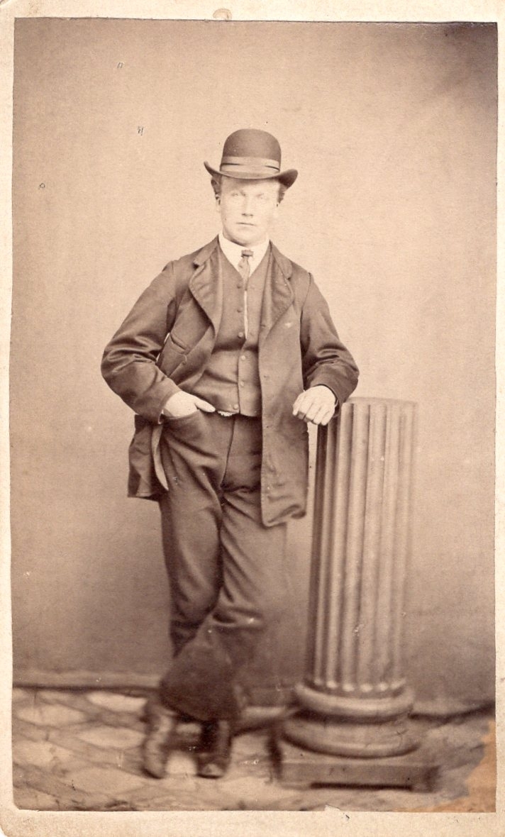 Ung mann stående i studio i Kragerø. Albuen på en søyle. Bowler hatt og dress. Ca. 1890 -1910