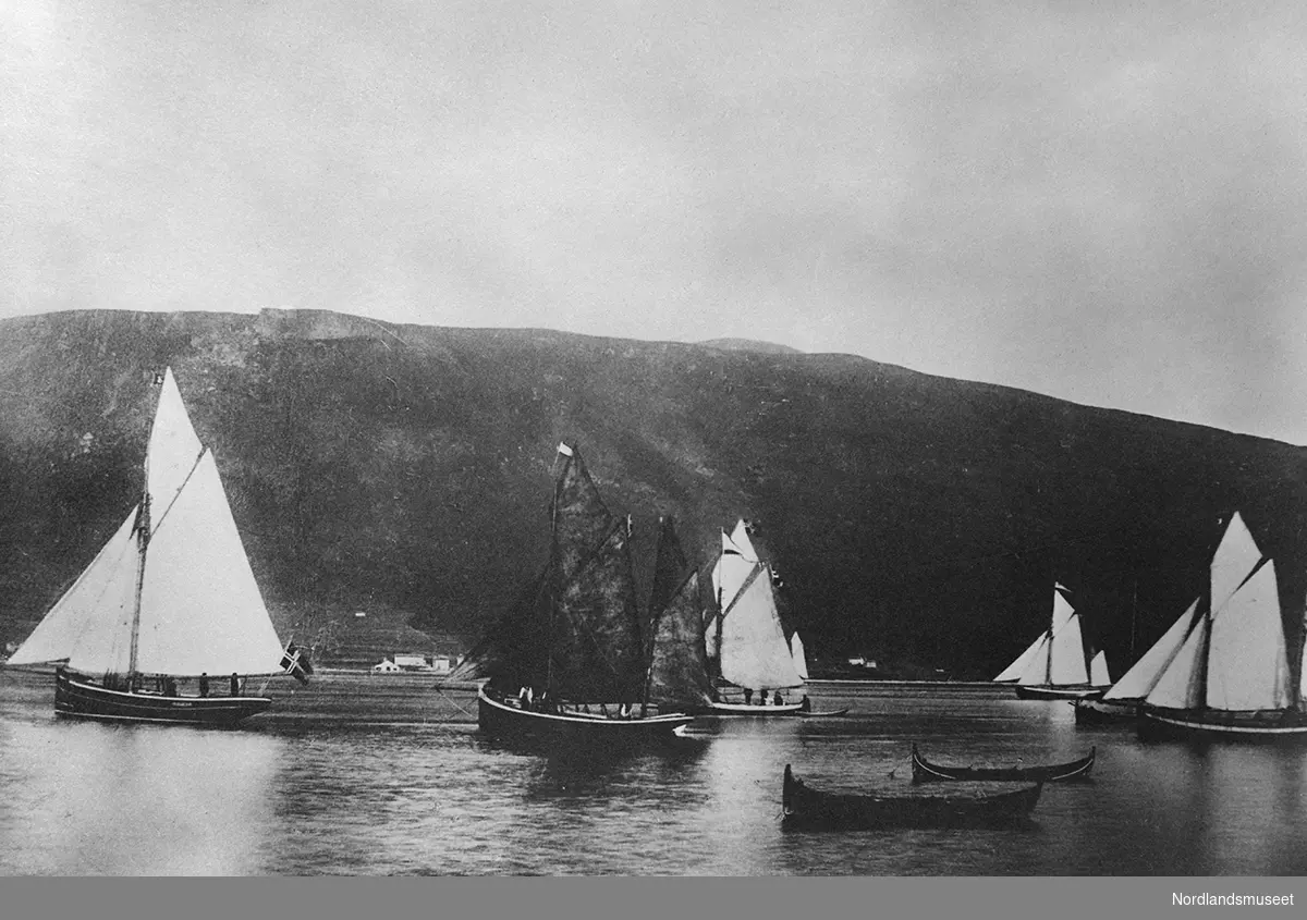 Kutter "Svanen" ytterst til venstre. Regattaen ved Fiskeriutstillingen i Tromsø 1894. Flere seilbåter med sneseil. To mindre nordlandsbåter i forgrunnen. Tilsammen syv båter, fem av dem under seil.
