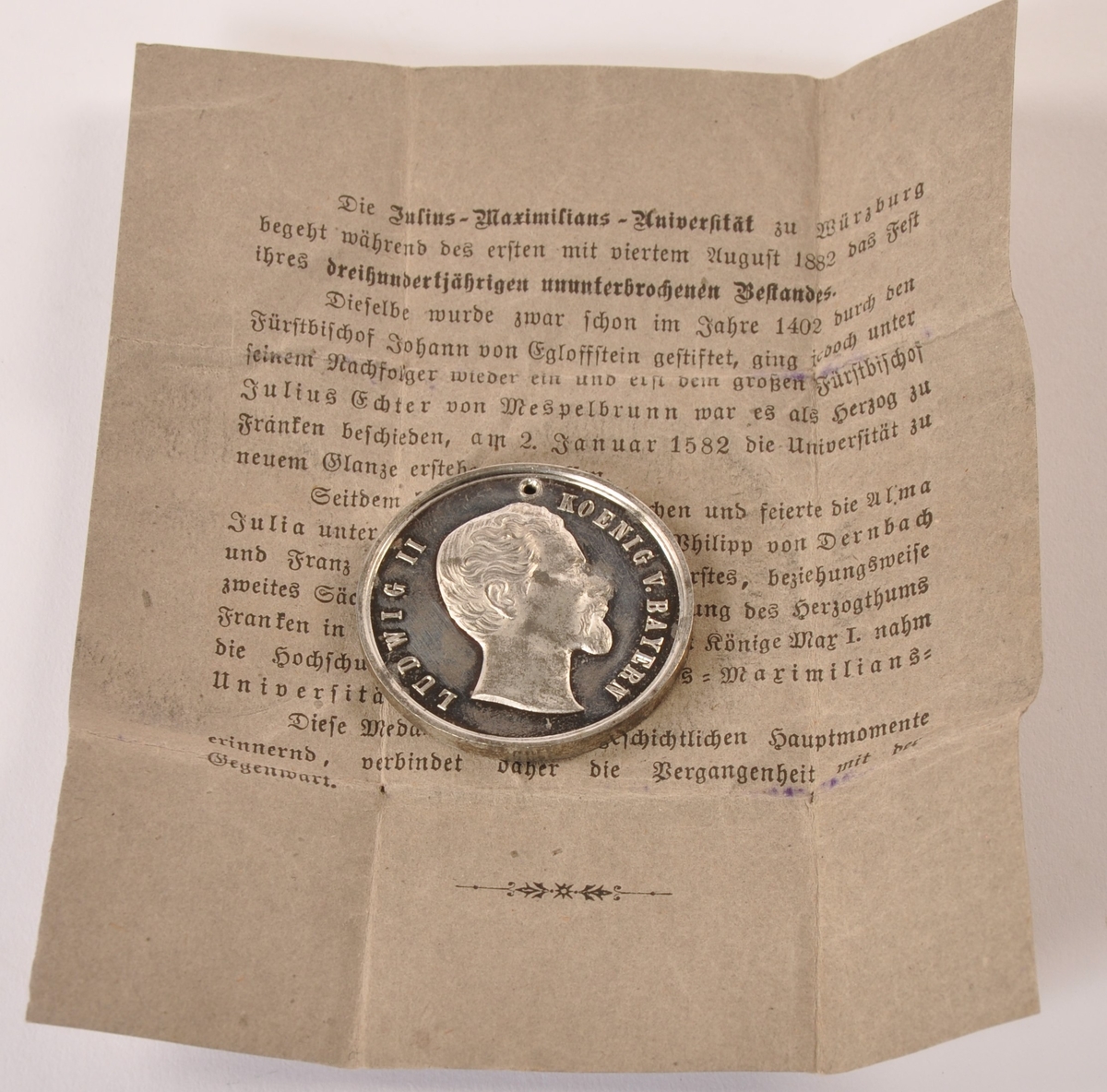 Sirkulær minnemedalje i metall, muligens sølv. Til minne om 300-års dagen av Julius Maximilians universtet i Würzburg. adversen: Portrett Ludvig av Bayern. Reversen: Våpenskjold med tekst. Ligger i brettet papirark med påtrykket forklaring av jubileet.