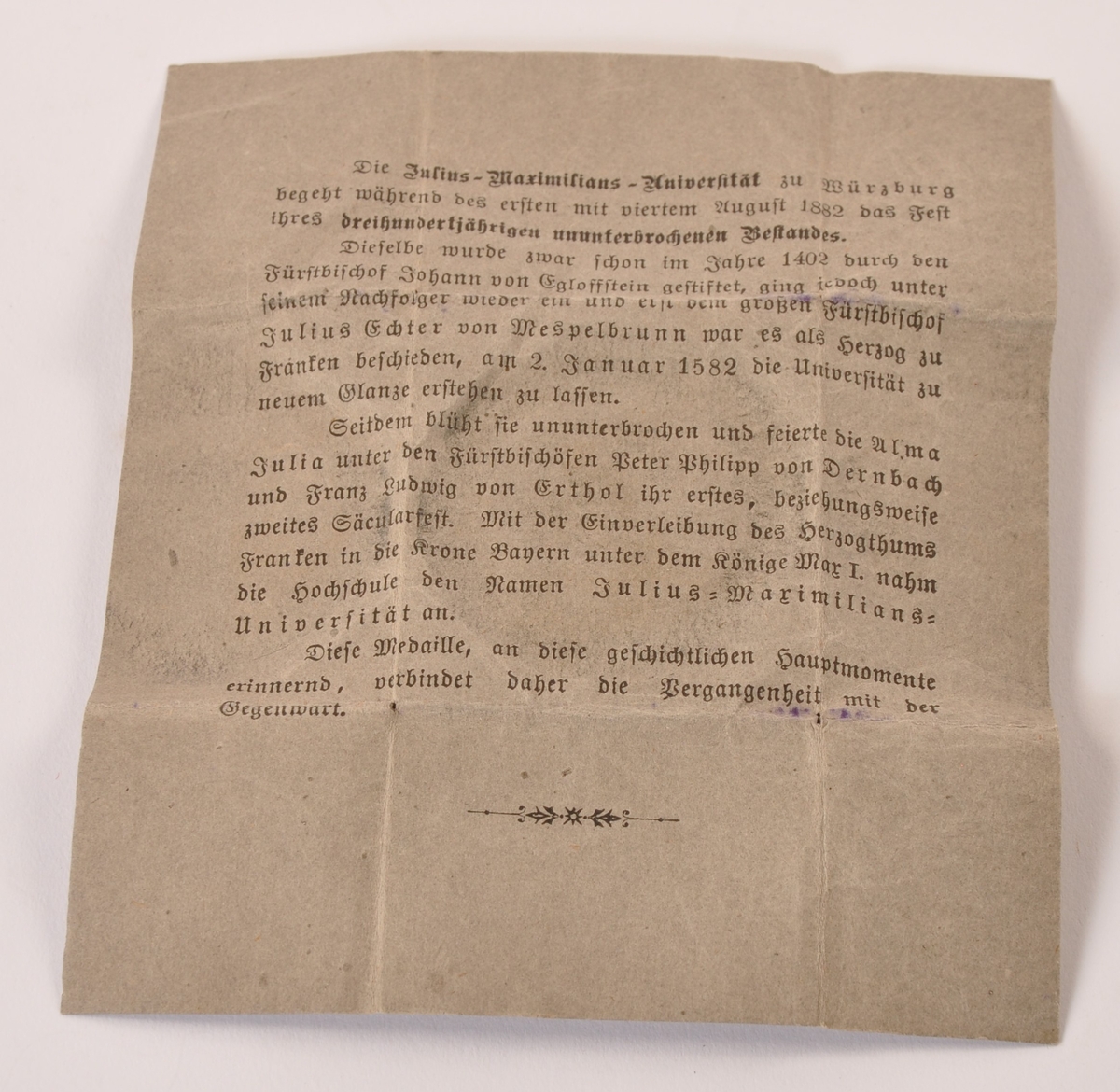 Sirkulær minnemedalje i metall, muligens sølv. Til minne om 300-års dagen av Julius Maximilians universtet i Würzburg. adversen: Portrett Ludvig av Bayern. Reversen: Våpenskjold med tekst. Ligger i brettet papirark med påtrykket forklaring av jubileet.