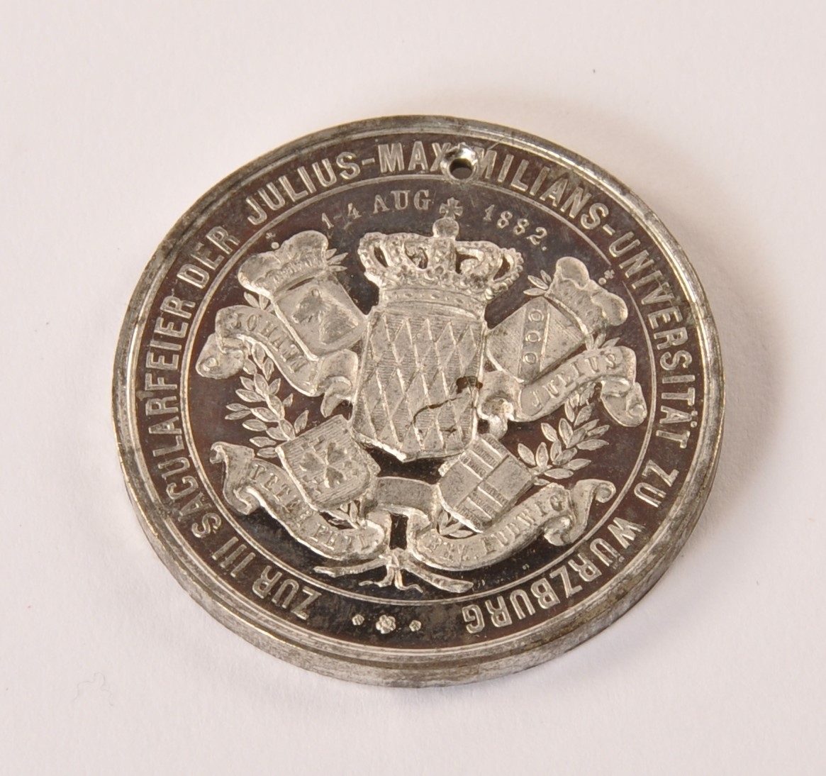 Sirkulær minnemedalje i metall, muligens sølv. Til minne om 300-års dagen av Julius Maximilians universtet i Würzburg. adversen: Portrett Ludvig av Bayern. Reversen: Våpenskjold med tekst. Ligger i brettet papirark med påtrykket forklaring av jubileet.