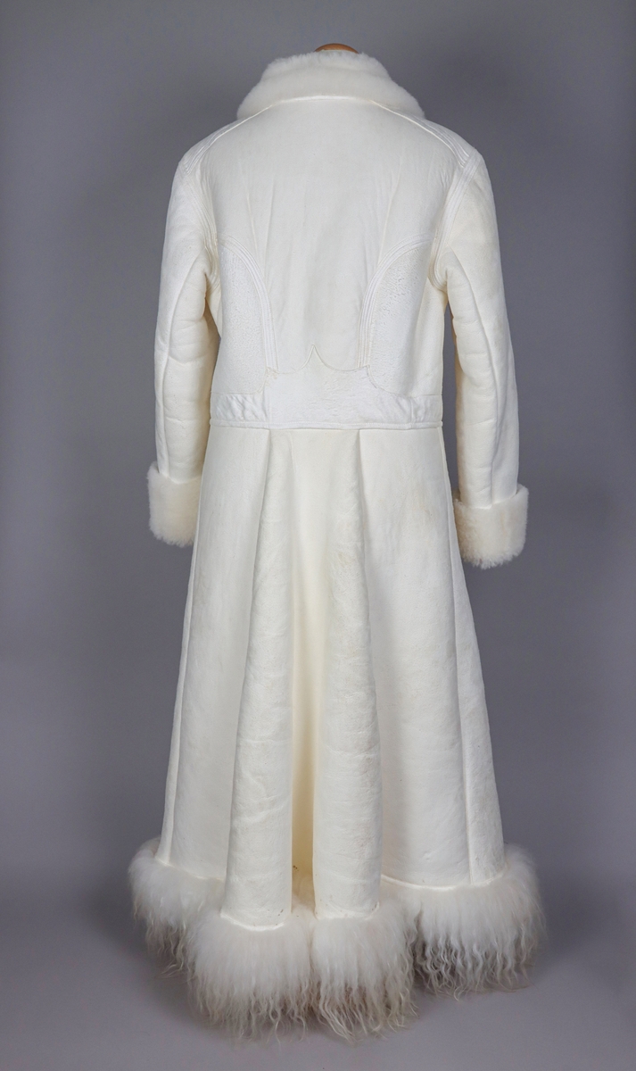 Hellång vit fårskinnspäls till mansdräkter från Västerdalarna. Pälssidan vänd innåt. Dubbelknäppning med 9 par mässingsknappar med sexuddiga stjärnorn på. Pälsen har långa pälslocksbräm längs neder- och framkanten, samt kortklippta uppvikta bräm vid ärmsluten.