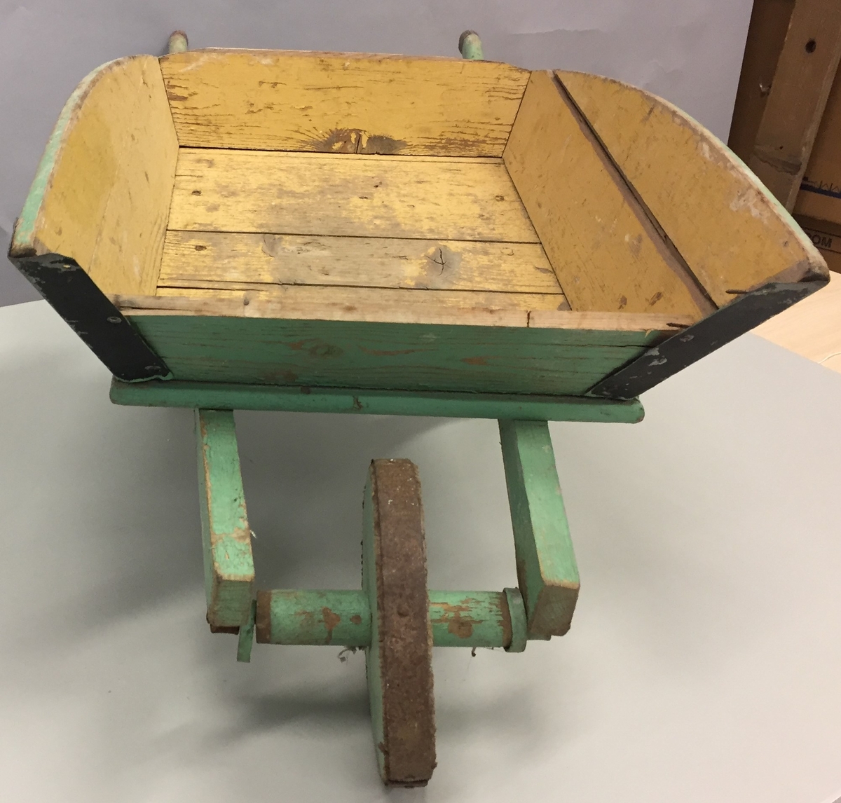 Trillebår i tre og metall. Truleg heimelaga. Trillebåra har eit hjul i tre, med ein ring ytterst av metall. Støtter slik at trillebåra kan stå støtt.To håndtak, Trillebåra er noko defekt.