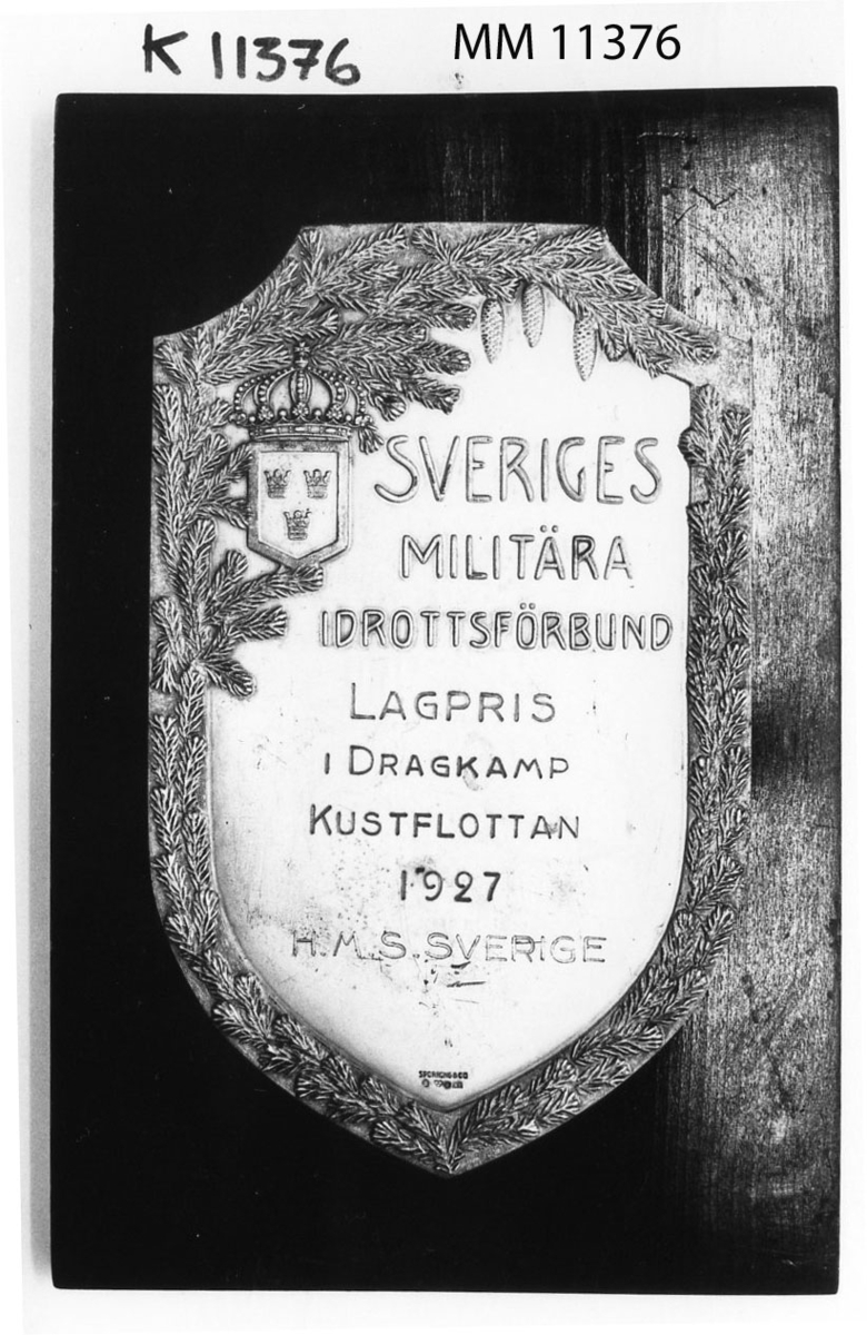 Plakett av silver på platta av trä, svartpolerad.
I relief Lilla Riksvapnet, granranka samt texten: Sveriges Militära Idrottsförbund. Märkta: Sporrong o Co samt silverstämpel.
Textad inskription: Lagpris i dragkamp Kustflottan 1927. H.M.S. Sverige.