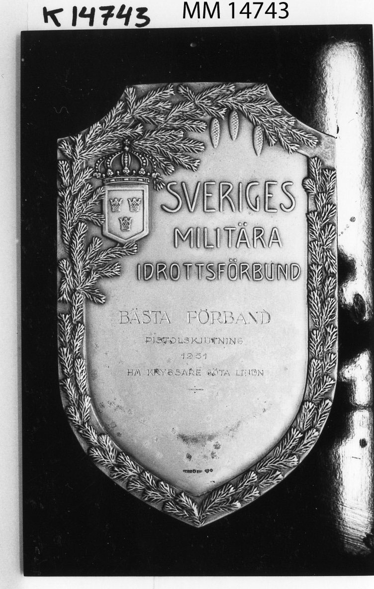 Plakett, idrottssköld i silver på platta av bakelit.
Text: Sveriges militära Idrottsförbund.
Ingravering: Bästa förband pistolskjutning 1951
HM Kryssaren Göta Lejon.