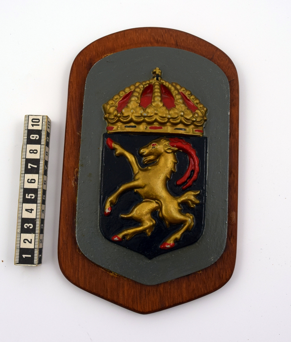 Vapensköld i metall på platta av trä. Motivet utgörs av jagaren Hälsinglands heraldiska vapen som föreställer en gyllene bock med röda horn i ett svart fält under kunglig krona.