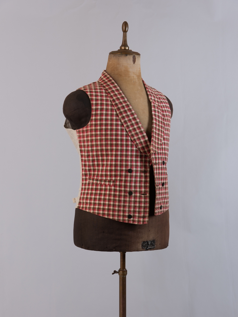 Vest i rutet bomullslerret i fargene rødt, olivengrønt/grønnnbrunt og ubleiket hvitt. Fôr og rygg i linlerret. Vesten er dobbeltspent med svarte, stofftrukne knapper. Den har sjalskrage og spensel i ryggen. Plagget er helt håndsydd. På forstykkene er det to lett skråstilte lommer og vesten buer ned midt foran.