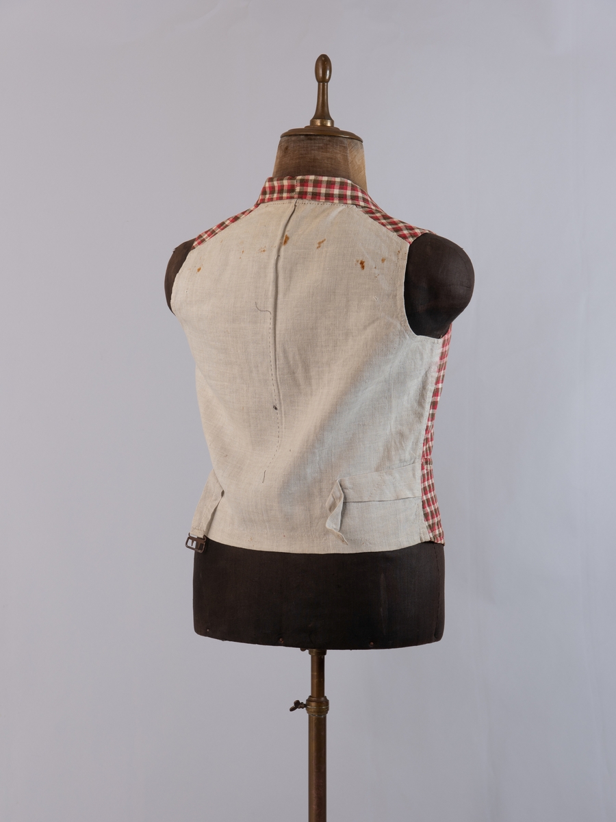 Vest i rutet bomullslerret i fargene rødt, olivengrønt/grønnnbrunt og ubleiket hvitt. Fôr og rygg i linlerret. Vesten er dobbeltspent med svarte, stofftrukne knapper. Den har sjalskrage og spensel i ryggen. Plagget er helt håndsydd. På forstykkene er det to lett skråstilte lommer og vesten buer ned midt foran.