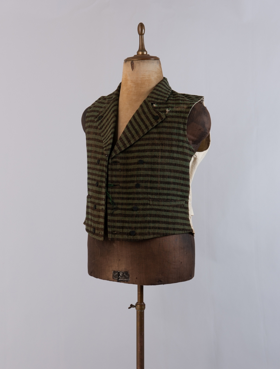 Dobbeltspent vest i ullverken, stripet i grønt og svart (sauesvart) med brun renning. Dobbel rygg i bomullslerret. Halvlinslerret (bomull/stry) i fôret i framstykkene. Vesten har fire knapphull sydd med kamgarnsullgarn og svarte knapper trukket med svart bomullssateng med brun renning. Plagget har nedbrettet krage og slag/reverser som kan kneppes mot bolen. Knapp og knapphull. Vesten buer ned midt framme og har svakt skråstilte lommer. Vattering av stry i framstykkene. I ryggen buer vesten opp midt bak. Den er vrangsydd langs ytterkantene og stukket ned med maskin, 0,8 cm fra kanten. (Registrering ved Bunad- og folkedraktrådet/Norsk institutt for bunad og folkedrakt v/Jon Fredrik Skauge, 2005, revidert v/Bjørn Sverre Hol Haugen, 2020.)