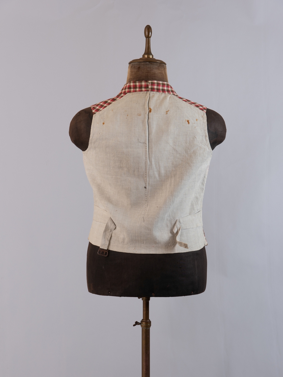 Vest i rutet bomullslerret i fargene rødt, olivengrønt/grønnnbrunt og ubleiket hvitt. Fôr og rygg i linlerret. Vesten er dobbeltspent med svarte, stofftrukne knapper. Den har sjalskrage og spensel i ryggen. Plagget er helt håndsydd. På forstykkene er det to lett skråstilte lommer og vesten buer ned midt foran.