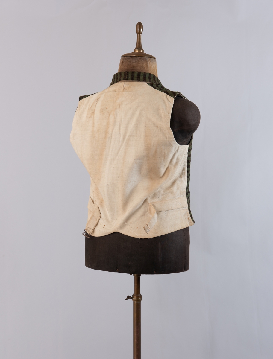 Dobbeltspent vest i ullverken, stripet i grønt og svart (sauesvart) med brun renning. Dobbel rygg i bomullslerret. Halvlinslerret (bomull/stry) i fôret i framstykkene. Vesten har fire knapphull sydd med kamgarnsullgarn og svarte knapper trukket med svart bomullssateng med brun renning. Plagget har nedbrettet krage og slag/reverser som kan kneppes mot bolen. Knapp og knapphull. Vesten buer ned midt framme og har svakt skråstilte lommer. Vattering av stry i framstykkene. I ryggen buer vesten opp midt bak. Den er vrangsydd langs ytterkantene og stukket ned med maskin, 0,8 cm fra kanten. (Registrering ved Bunad- og folkedraktrådet/Norsk institutt for bunad og folkedrakt v/Jon Fredrik Skauge, 2005, revidert v/Bjørn Sverre Hol Haugen, 2020.)