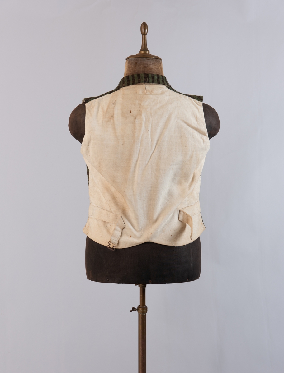 Dobbeltspent vest i ullverken, stripet i grønt og svart (sauesvart) med brun renning. Dobbel rygg i bomullslerret. Halvlinslerret (bomull/stry) i fôret i framstykkene. Vesten har fire knapphull sydd med kamgarnsullgarn og svarte knapper trukket med svart bomullssateng med brun renning. Plagget har nedbrettet krage og slag/reverser som kan kneppes mot bolen. Knapp og knapphull. Vesten buer ned midt framme og har svakt skråstilte lommer. Vattering av stry i framstykkene. I ryggen buer vesten opp midt bak. Den er vrangsydd langs ytterkantene og stukket ned med maskin, 0,8 cm fra kanten. (Registrering ved Bunad- og folkedraktrådet/Norsk institutt for bunad og folkedrakt v/Jon Fredrik Skauge, 2005, revidert v/Bjørn Sverre Hol Haugen, 2020.)